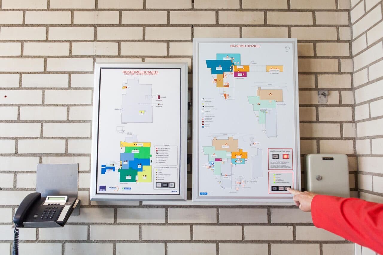 Ontruimingsplattegronden naast een brandmeldpaneel, iemand wijst een vluchtroute aan