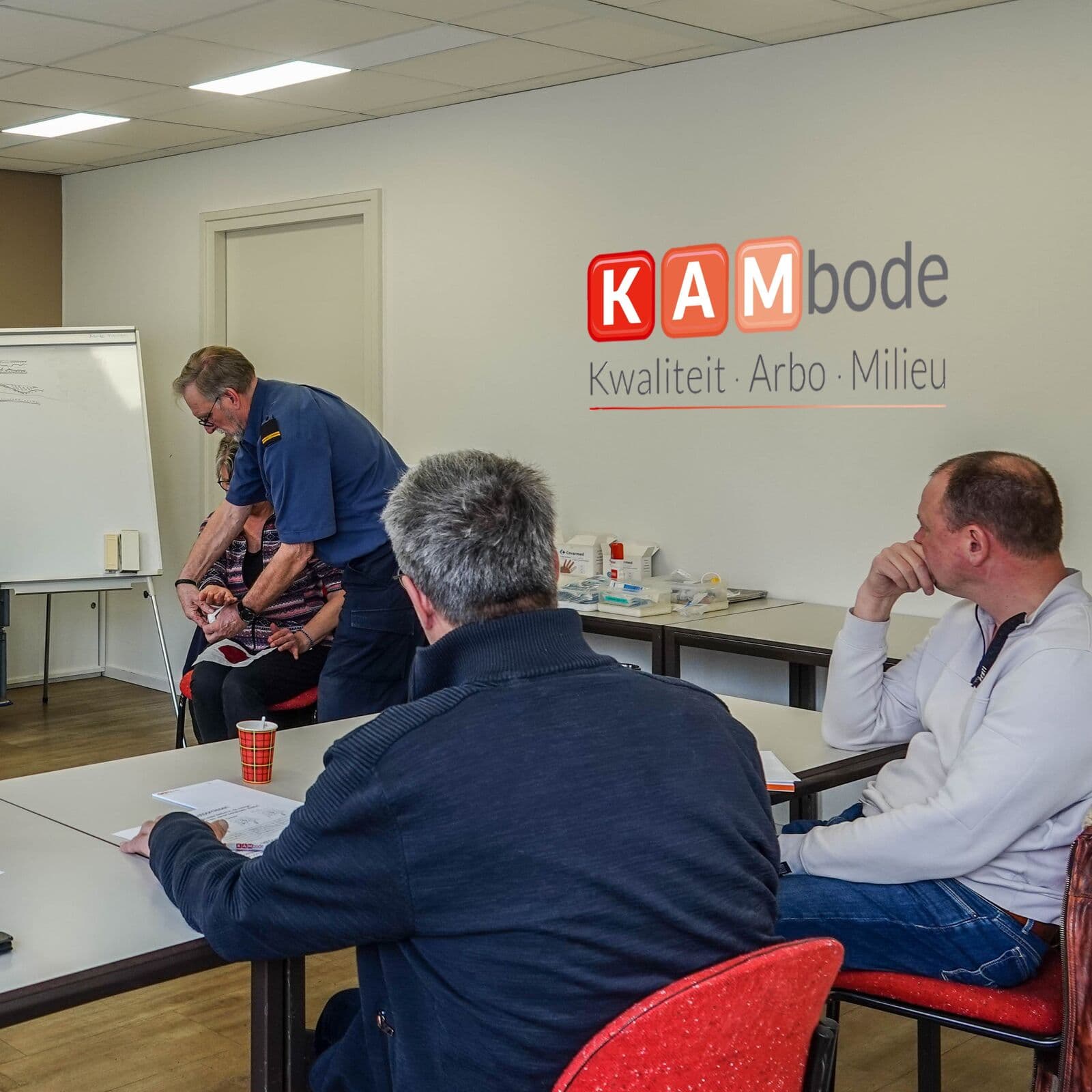 Docent van KAMbode geeft een praktijkles aan een groep cursisten, met het KAMbode-logo op de muur